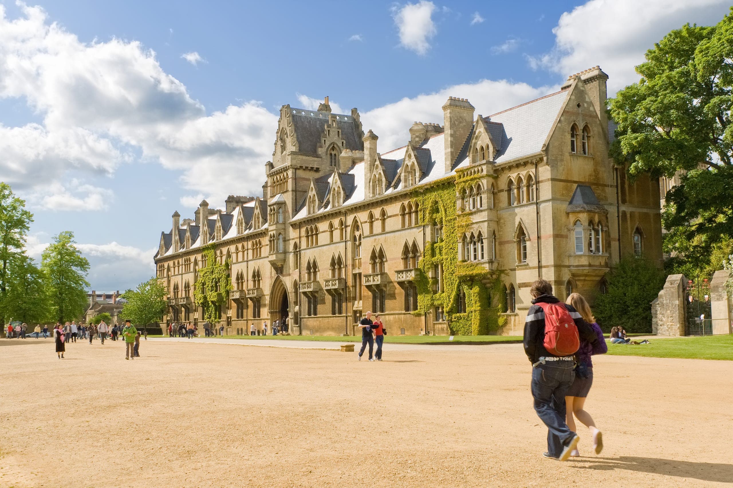 Several individuals stroll past a prominent university building, showcasing its architectural features against a bright sky.